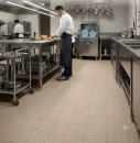 Nowoczesna kuchnia restauracyjna ze stanowiskami ze stali nierdzewnej, w której kucharz przygotowuje posiłki. Na podłodze ułożone są beżowe, matowe płytki gresowe Marazzi SistemT - Graniti Panna Diamond 20x20 cm M65G.