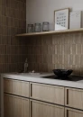 Gayafores Deco Kirei Caramel 32x62,5- brązowa lamelowana płytka ścienna w połysku wyłożone na rękawie kuchennym w zestawieniu z brązową drewnianą zabudową kuchenną oraz jasnym kamiennym blatem. Drewniana półka na produkty, czarna płyta kuchenna.