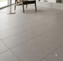 Zdjęcie ukazuje podłogę z kwadratowych płytek Marazzi SistemT Graniti Grigio Medio Diamond 20x20 cm M7LL. Jasnoszare, matowe kafle o strukturze granitu ułożono w równą siatkę w nowoczesnym, jasnym wnętrzu z fotelem w tle.