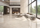 ITT Taj Mahal Polished 120x120- gresowa płytka imitująca beżowy klasyczny kamień z przebarwieniami z dużym formacie i połysku na podłodze i ścianach przestronnego, oświetlonego salonu z antresolą. Przeszklona ściana w czarnych ramach.