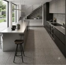 Nowoczesna kuchnia industrialna, w której na podłodze leżą szare, groszkowane płytki Marazzi SistemT - Graniti Grigio Medio Antislip R11 20x20 cm M7LE. Widać stalową wyspę, ciemną zabudowę i schody, a całość doświetla duże okno tarasowe.