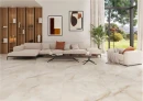 Keratile Orion Cream Satin 100x100 Gresowa płytka podłogowa imitująca kamień onyks w beżowym/kremowym kolorze i jasnymi oraz brązowymi żyłami na podłodze otwartego, przestronnego salonu. Białe kanapy na czarnych nogach, drewniany regał ścienny.