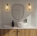 Wnętrze łazienki ze ścianą z drobnej, białej mozaiki (płytki WOW 3DB Bevel Quartz 12,5x25 cm 135546). Na drewnianej szafce stoi biała umywalka nablatowa i czarny dozownik. Nad nimi wisi organiczne lustro w czarnej ramie, flankowane przez dwa kinkiety.