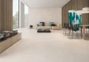 Prissmacer Cross Easton Marfil Mat 120x120- wielkoformatowa płytka gresowa imitująca beton w szarym kolorze o ciepłym odcieniu na podłodze i ścianie salonu w połączeniu z drewnianą lamelą na ścianach. Beżowa kanapa, ciemny kamienny stolik kawowy.