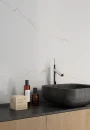 Prissmacer LAB. Bianco Mare Gold Lapp 120x120- biały marmur ze złotymi żyłami i wykończeniu lapatto na ścianie łazienki. Czarny blat ustawiony na drewnianej szafce, kamienna grafitowa umywalka wolnostojąca, chromowana nowoczesna bateria umywalkowa.