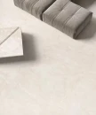 Ergon Matera Stone Neutra White Silktech Rett. 120x120 EN6C- gresowa płytka imitująca jasny szary kamień na podłodze salonu zestawiona z szarą kanapą oraz kamiennym geometrycznym stolikiem kawowym.