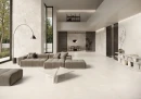 Ergon Matera Stone Neutra White Silktech 60x120 EN52- gresowa płytka imitująca minimalistyczny szary jasny kamień z przebarwieniami na podłdoze przestronnego salonu. Szara kanapa, kamienny marmurowy stolik z marmuru, duże okna w czarnej ramie.