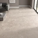 Casalgrande Padana Pietra Tiburtina Campidoglio Cross 120x120- gresowa płytka podłogowa imitująca beżowy trawertyn z szarymi przebarwieniami położona na podłodze w salonie gdzie znajduje się szary wypoczynek oraz duże okno