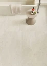 Emil/Provenza Unique Travertine Minimal White Lappato Antique Rett. 120x120 ENLP beżowo-szara gresowa płytka imitująca kamień z jasnymi przebarwieniami  na podłodze łazienki. Przyścienna geometryczna wanna, nowoczesny stolik w bezowym kolorze.