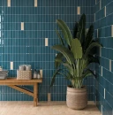 Ściana ułożona z pionowych, ciemnoniebieskich błyszczących płytek WOW Casablanca Azure Gloss 5x15 135380 z beżowymi akcentami. Przy ścianie stoi drewniana ławka z ręcznikami oraz duża roślina doniczkowa w plecionym koszu.