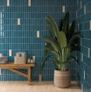 Aranżacja wnętrza ze ścianą wyłożoną pionowo błyszczącymi, ciemnoniebieskimi płytkami WOW Casablanca O Azure Gloss 5x15 135388 cm. Obok ściany stoi duża roślina w plecionym koszu oraz drewniana ławka z ręcznikami.