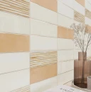 Ściana wyłożona błyszczącymi płytkami strukturalnymi i gładkimi WOW Casablanca Comb Lime Gloss 5x15 cm 135391 w kolorach kremowym i pomarańczowym. Układ cegiełki. W rogu wazon z suszonymi kwiatami i książka.