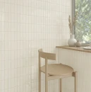 Jasny, minimalistyczny kącik ze ścianą z pionowych, błyszczących płytek WOW Colour Notes Oat 4x12,5 132971. Geometryczny, jasny drewniany hoker stoi przy blacie z wazonami i suszonymi trawami przed oknem z widokiem na zieleń.