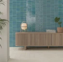 Eleganckie wnętrze ze ścianą wyłożoną niebieskimi, błyszczącymi płytkami WOW Colour Bars Azur 4x12,5 cm (133160). Przed ścianą stoi drewniana ryflowana komoda, na której ustawiono lampę stołową, książki oraz roślinę doniczkową.