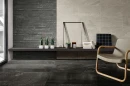 Coem Blendstone Graphite Lucidato 60x120 BL627LR grafitowa płytka gresowa imitująca kamień z jasnymi przebarwieniami na podłodze salonu w zestawieniu z beżową płytką na ścianie. Jasne krzesło z drewnianą ramą i beżowym obiciem
