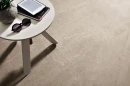 Coem Blendstone Beige Naturale 60x60 BL602R gresowa płytka w beżowym kolorze imitująca kamień z jasnymi przebarwieniami na podłodze salonu. Biały stolik kawowy na trzech nogach. Książka i czytnik eboków na blacie.