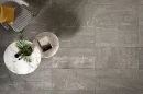 Coem Blendstone Dark Grey Naturale 60x60 BL600R gresowa płytka imitująca ciemny szary kamień z jasnymi żyłami i przebarwieniami na podłodze salonu. Okrągłe stoliki z białym marmurowym blatem, fotel z obszyciem w biało czarną kratkę.