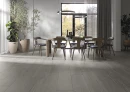 Jadalnia z drewnianym stołem, drewnianymi krzesłami z czarnym skórzanym obiciem i płytkami podłogowymi szarymi drewnopodobnymi Cifre Adena Grey Mate XS 30x120