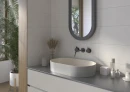 Jasna, biała łazienka z płytkami ściennymi imitującymi beton Cifre Alure White Mate Xs Pb 25x50 XS PB, owalną nablatowa umywalką, grafitowym podwieszanym blatem, czarną ścienną armaturą, zaokrąglonym lustrem w ciemnej ramie.