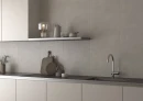 Minimalistyczna kuchnia z szarymi płytkami z imitacji betonu Cifre Alure Plomb Mate Xs Pb 25x50 XS PB, z jasnobeżowymi frontami, ciemnoszarym blatem, podblatowym zlewozmywakiem, chromowaną baterią kuchenną, otwartą półką i dekoracyjną lampą.