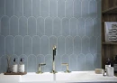 Ściana z niebieskimi płytkami ściennymi, imitującymi beton w kształcie rybiej łuski Cifre Alure Scale Aqua Mate 8x21,5, z lśniącą złotą armaturą, drewnianą otwartą szafką w kolorze ciemnego drewna, drewnianą tacą na kosmetyki i świeczką.