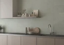 Ściana w kuchni z zielonymi płytkami ściennymi, imitującymi beton, pochodzenia hiszpańskiego Cifre Alure Sage Mate Xs Pb 25x50 XS PB, z ciemnym blatem, frontami w kolorze drewna, dekoracyjną półką z dodatkami, drewnianymi i pastelowymi dodatkami.