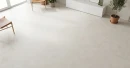 Biała podłoga z płytkami hiszpańskimi, antypoślizgowymi, imitującymi beton   Cifre Alure White Mate Sl Rc Antislip 60x120 SL RC, z białą szafką RTV, drewnianymi krzesłami z siedziskiem ze skóry, żywymi kwiatami w doniczkach.