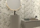Jasna ściana z dekoracyjnymi płytkami, z motywem liści eukaliptusa Cifre Alure Lipra Ivory Mate Xs Pb 25x50 XS PB, z podłużną jasną umywalką, czarną armaturą, podświetlonym lustrem, wiszącą jasną szafką i stonowanymi dodatkami.