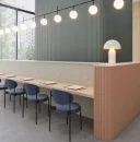 Scena restauracyjna z długim stołem i niebieskimi krzesłami, oświetlona kulistymi lampami. W tle ściana wyłożona ryflowanymi płytkami WOW Faces Dune Steel Blue 5x40 131488, obok jasny pas i brzoskwiniowa lada.