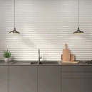 Nowoczesna kuchnia z szarymi meblami i zlewem. Ściana nad blatem wyłożona białymi, błyszczącymi płytkami 3D WOW Faces Dune White Gloss 5x40 131798 o strukturze fali. Dwie industrialne lampy wiszące, deski do krojenia i zioła w doniczce.