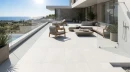 Taras z płytkami antypoślizgowymi, białymi imitującymi beton Cifre Acron White Mate Rc 20mm Antislip 60x120, ze szklaną balustradą, strefą prysznicową, wypoczynkiem w kolorze jasnym z drewnianymi dodatkami oraz stolikiem kawowym.