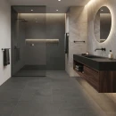 Ciemna łazienka z płytkami antypoślizgowymi, imitującymi beton Cifre Acron Anthracite Mate Antislip 90x90 z podwieszaną szafką umywalkową z  drewna z czarnym blatem, oświetleniem liniowym, okrągłym lustrem oraz kabiną walk-in z czarnymi profilami.
