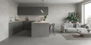 Salon połączony z kuchnią z płytkami imitującymi beton, Cifre Acron Pearl Mate N-Plus SI Rect. 30x60 z aneksem z matowych, ciemnoszarych szafek stojących oraz wiszących, jasnoszarą sofą, niskim stolikiem kawowym, dywanem.