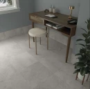 Gresowe płytki podłogowe i cokołowe WOW Halo XXL Noon 44x44 (132941) imitujące szary beton lub kamień w nowoczesnym gabinecie z drewnianym biurkiem, stołkiem i rośliną.