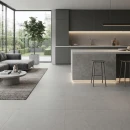 Salon z aneksem kuchennym z płytkami wielkoformatowymi imitującymi beton Cifre Acron Pearl Mate N-plus 120x120, z ciemnoszarymi, matowymi szafkami, wyspą kuchenną z podświetleniem LED, szarą sofą, niski betonowym stolikiem, narożnym oknem tarasowym.