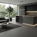 Salon z kuchnią z ciemnymi płytkami, antypoślizgowymi imitującymi beton Cifre Acron Anthracite Mate Antislip 120x120, z aneksem z ciemnych, matowych szafek, wyspy kuchennej, podświetleniem LED, ciemną sofą, dywanem, niskim betonowym stolikiem kawowym.
