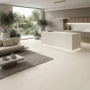 Salon z kuchnią z płytkami antypoślizgowymi, imitującymi beton, beżowymi Cifre Acron Ivory Mate Antislip 120x120, z podświetlaną wyspą kuchenną, beżową sofą, kamiennym stolikiem, złotym oświetleniem, roślinami w donicach.