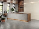 Recepcja z płytkami ściennymi imitującymi beton, pochodzenia hiszpańskiego Cifre Acron Ivory Mate Xs Rect. 40x120, z masywną ladą recepcyjną z drewnianym wykończeniem, dużymi oknami w czarnej ramie, skórzanymi fotelami.