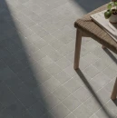 Zbliżenie na podłogę wyłożoną szarymi płytkami WOW Halo S Noon 11x11 (132946) w równym układzie siatkowym. Matowe gresy o strukturze kamienia z drobinami kruszywa oświetlone naturalnym światłem. Obok stoi drewniany stołek ze plecionym siedziskiem.