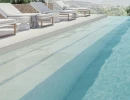 Elegancka strefa basenowa na zewnątrz wyłożona beżowymi, antypoślizgowymi płytkami WOW Marble Sand R11 (132924) na brzegu i schodach. Obok basenu stoją białe leżaki tarasowe z widokiem na górzysty krajobraz.