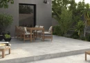 Taras z płytkami imitującymi kamień Cifre Arven Almond Mate Sl Rc N-Plus 60x120 SL RC, z drewnianym stołem i krzesłami, czarnym kinkietem, bujną roślinnością, czarnymi dodatkami.